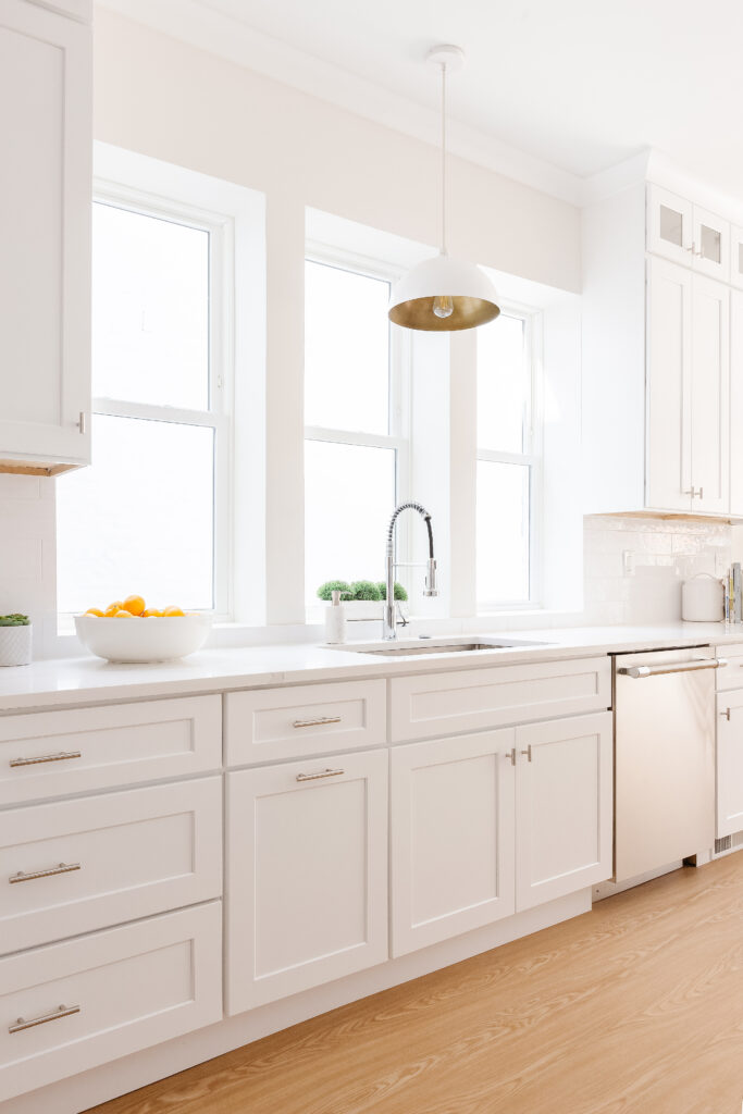 One side of a galley style kitchen features white cabinets, a large sink, stainless steel dishwasher and a marble counter top.